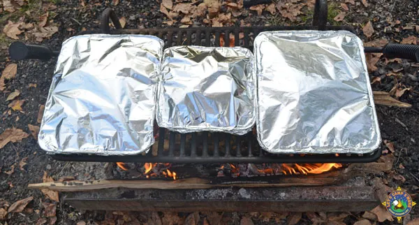 3 foil pans cooking on a grate over a campfire