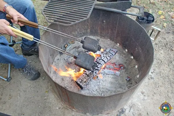 man holding 2 pie irons over a campfire ring