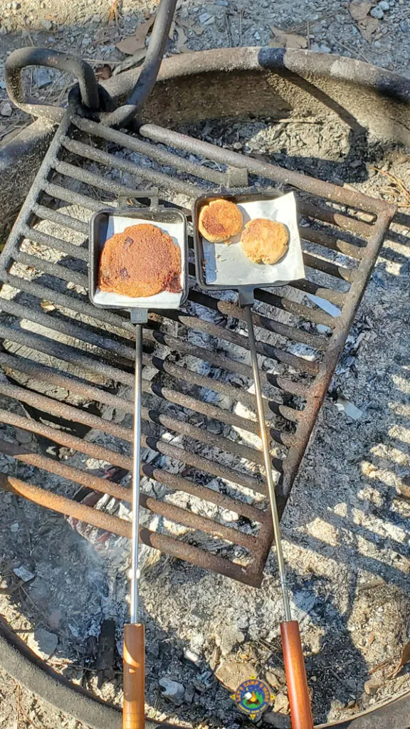 3 camping cookies in pie irons on top of a grate over a campfire