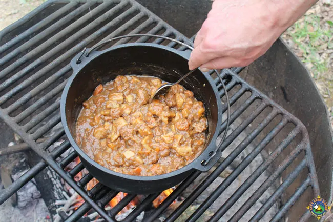 stew being stirred in a Dutch oven on a grate over a campfire