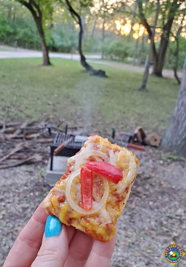 close up of a piece of camping pizza being held up at a campground with a campfire and sunset in the background