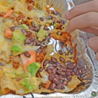 close up of a hand reaching for a cheesy chip from nachos made in a foil pan
